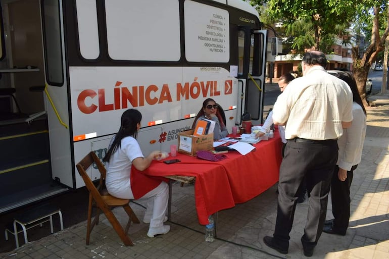 Dos mujeres en un escritorio con mantel rojo y un hombre hablando, rodeados por el ambiente de una clínica móvil.