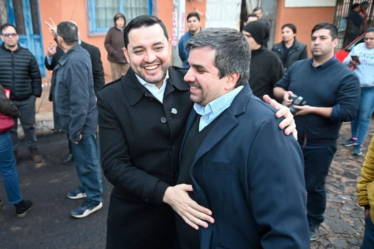 Luis Bello con abrigo negro y Derlis Bobadilla en traje azul, abrazándose sonrientes, rodeados de un grupo diverso de personas en un ambiente festivo.