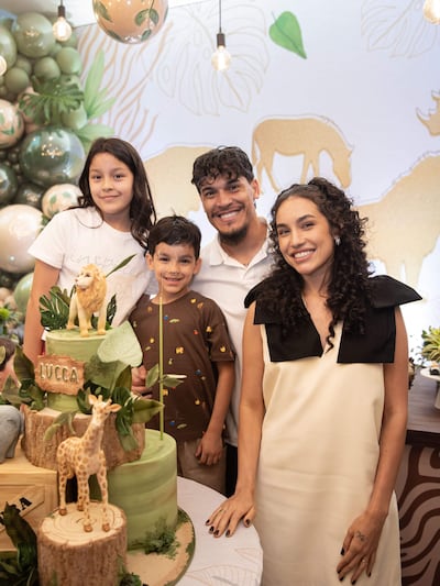 Niña con camiseta blanca y sonrisa, niño en primer plano con camiseta marrón, hombre con cabello rizado y mujer con vestido blanco, todos felices en una fiesta.