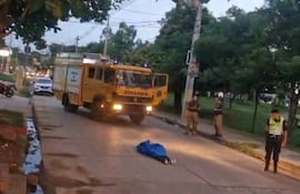 Cuerpo cubierto con manta azul en la calzada, camión de bomberos amarillo cercano y policías observando la situación.