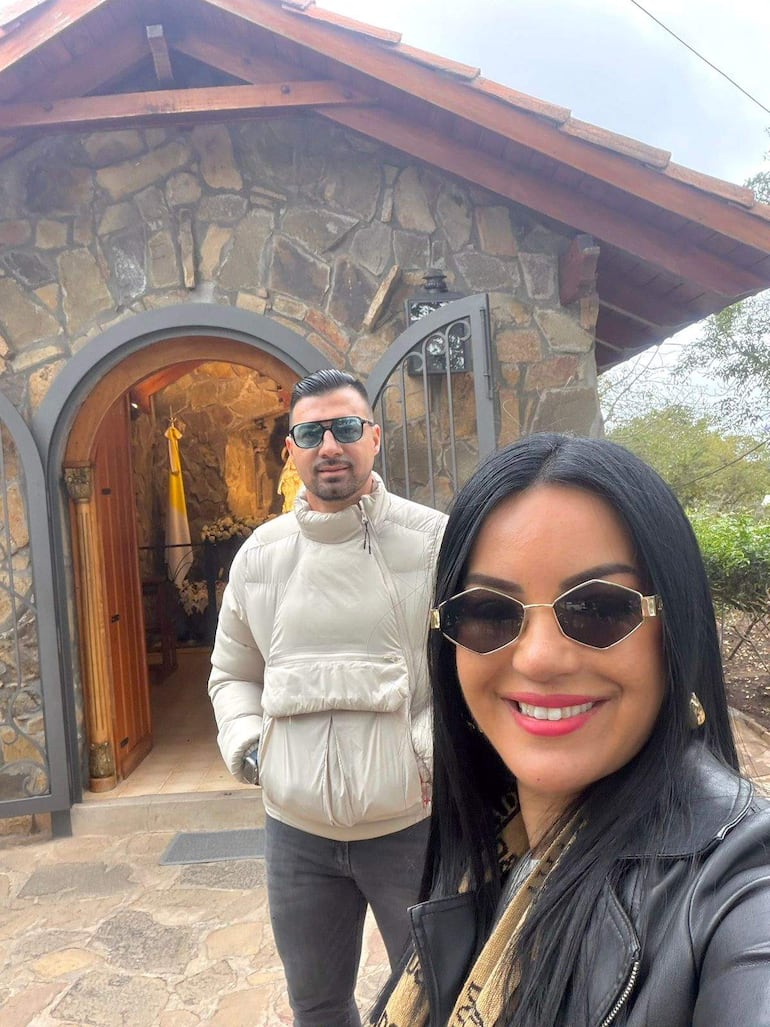 Julio César Cáceres con chaqueta blanca y gafas de sol, junto a Gloria Vera en chaqueta negra, ambos sonriendo en un entorno rústico.