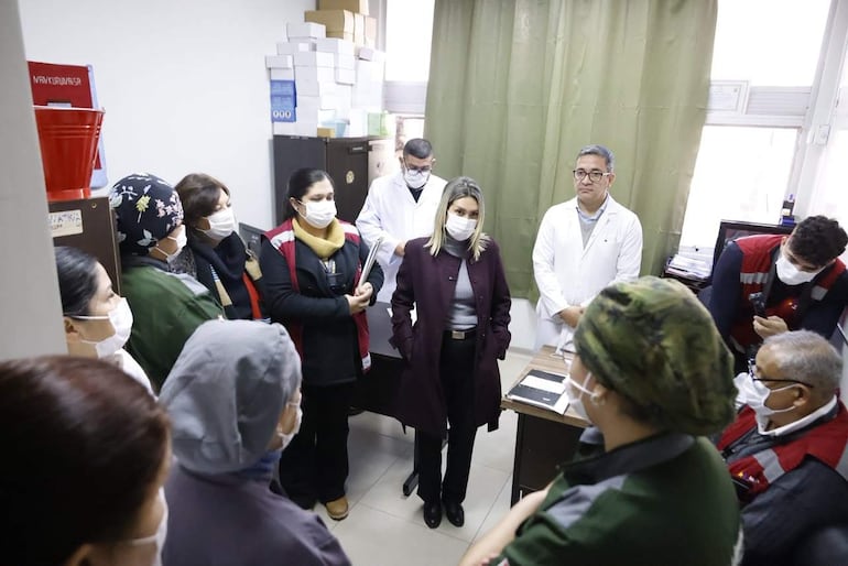 Mónica Recalde con abrigo oscuro en el centro, rodeada de personas con mascarillas y profesionales de la salud en blusas blancas.