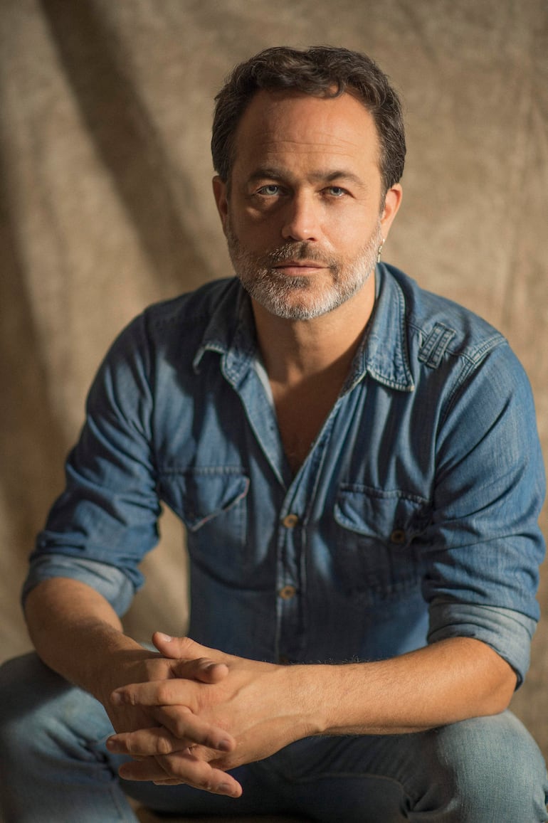 Gastón Pauls, con barba y cabello castaño, viste camisa de mezclilla y jeans, mientras mira directo a la cámara.