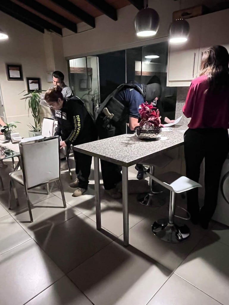 Mujer con cabello recogido sentada, dos hombres de pie revisan documentos, otra mujer observa en ambiente interior con luz tenue.