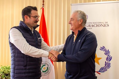 Gustavo Alfaro sonríe mientras estrecha la mano de un hombre en abrigo azul, frente a la bandera de Paraguay y un cartel de Migraciones.