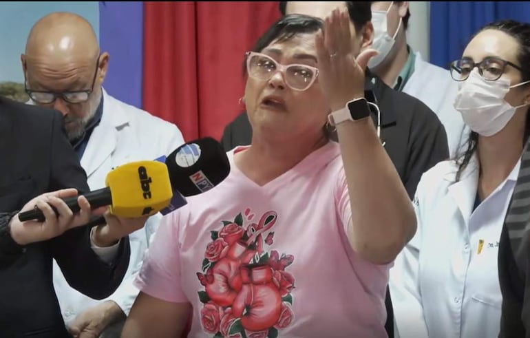 María Estela Galeano con polo rosa, gesticulando apasionadamente, mientras dos médicos en bata blanca la escuchan atentamente en un ambiente de conferencia.