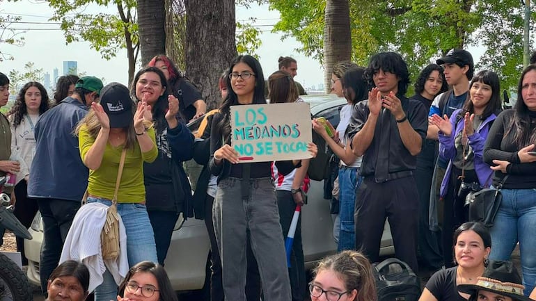 Mujer con gafas sostiene cartel 'LOS MEDANOS NO SE TOCAN', rodeada de personas aplaudiendo en un ambiente urbano.