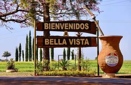 Cartel de madera rústico que dice 'BIENVENIDOS A BELLA VISTA', acompañado de una jarra grande, con campo verde y árboles al fondo.