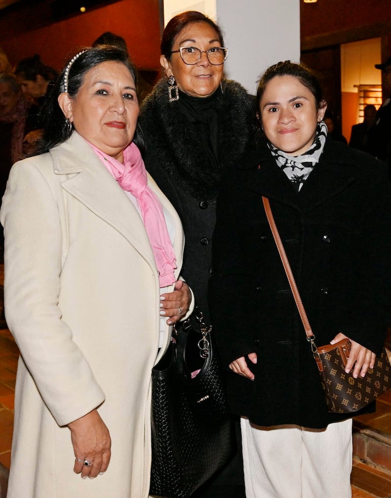 Tres mujeres de pie frente a una pared; una en abrigo blanco y bufanda rosa, otra en abrigo negro, y otra con bolso distintivo.