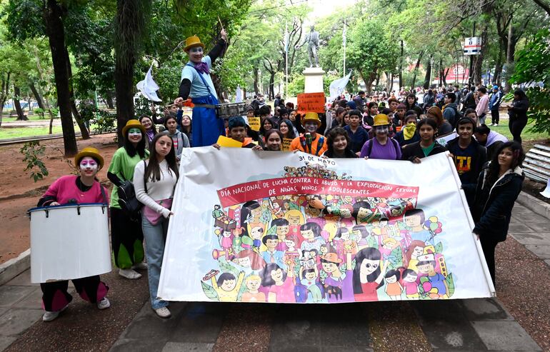 Grupo de jóvenes con atuendos coloridos en manifestación, dos payasos y mujer con cartel sobre abuso sexual infantil.