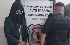Hombre con saco oscuro y camiseta blanca, con head cover negro que dice 'POLICÍA NACIONAL', frente a un policía en uniforme táctico.
