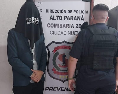 Hombre con saco oscuro y camiseta blanca, con head cover negro que dice 'POLICÍA NACIONAL', frente a un policía en uniforme táctico.