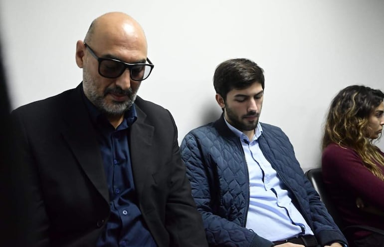 Benjamín Zapag, con chaqueta gris, y su padre Raúl, en traje negro, lucen serios en un entorno sobrio y iluminado con tonos grises.