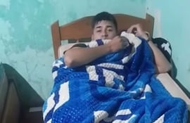 Joven policía acostado en cama, envuelto en manta azul en habitación con pared verde desgastada.