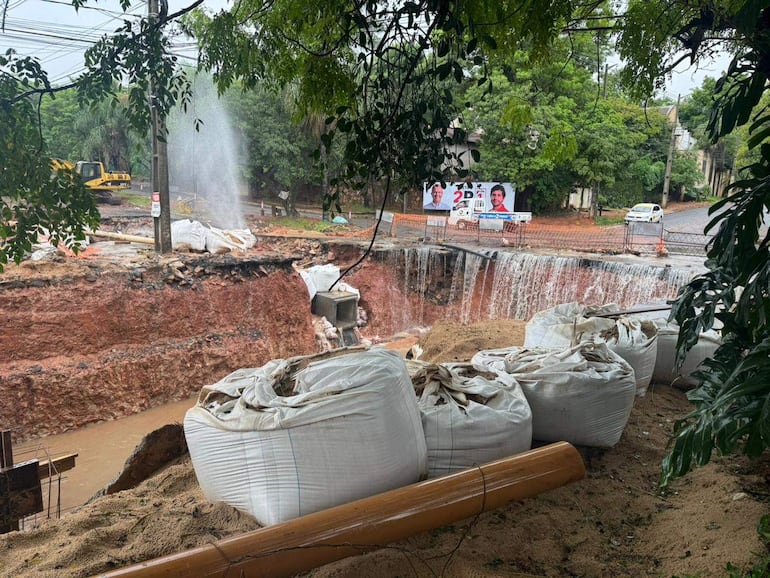 Zona de obra con zanja llena de agua y sacos de construcción apilados, maquinaria y poste de electricidad de fondo.