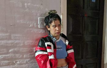 Mujer de pie con chaqueta roja y negra, camiseta azul claro, expresión seria, apoyada contra una pared de ladrillos blancos.
