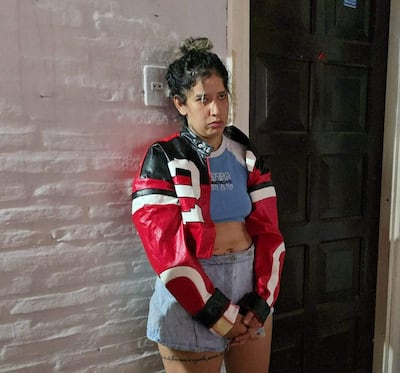 Mujer de pie con chaqueta roja y negra, camiseta azul claro, expresión seria, apoyada contra una pared de ladrillos blancos.