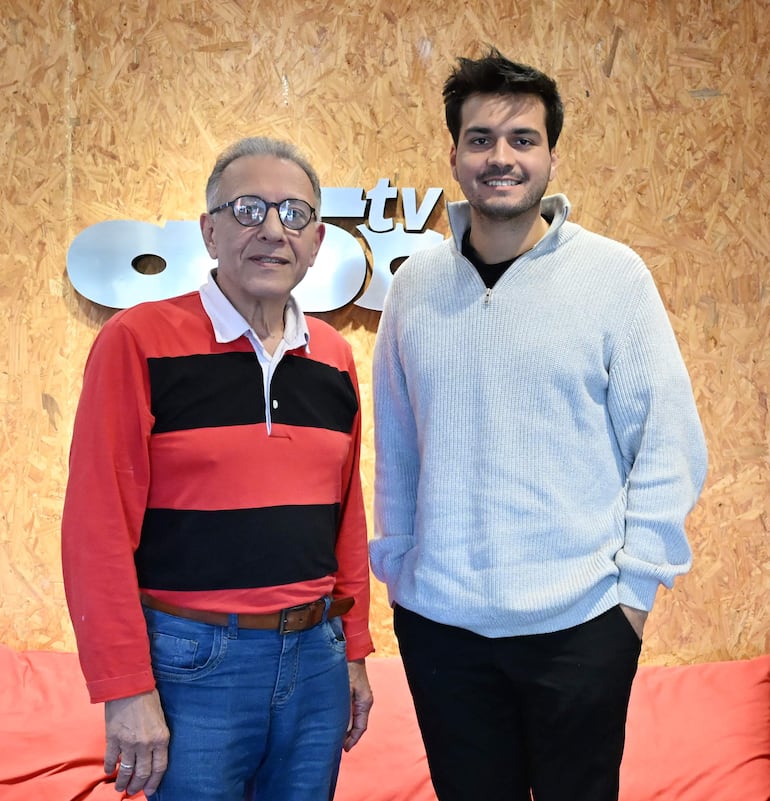 Hombre canoso en camiseta roja y pantalón de mezclilla, junto a hombre joven en suéter gris, sonriendo en un fondo con logo de abc tv.