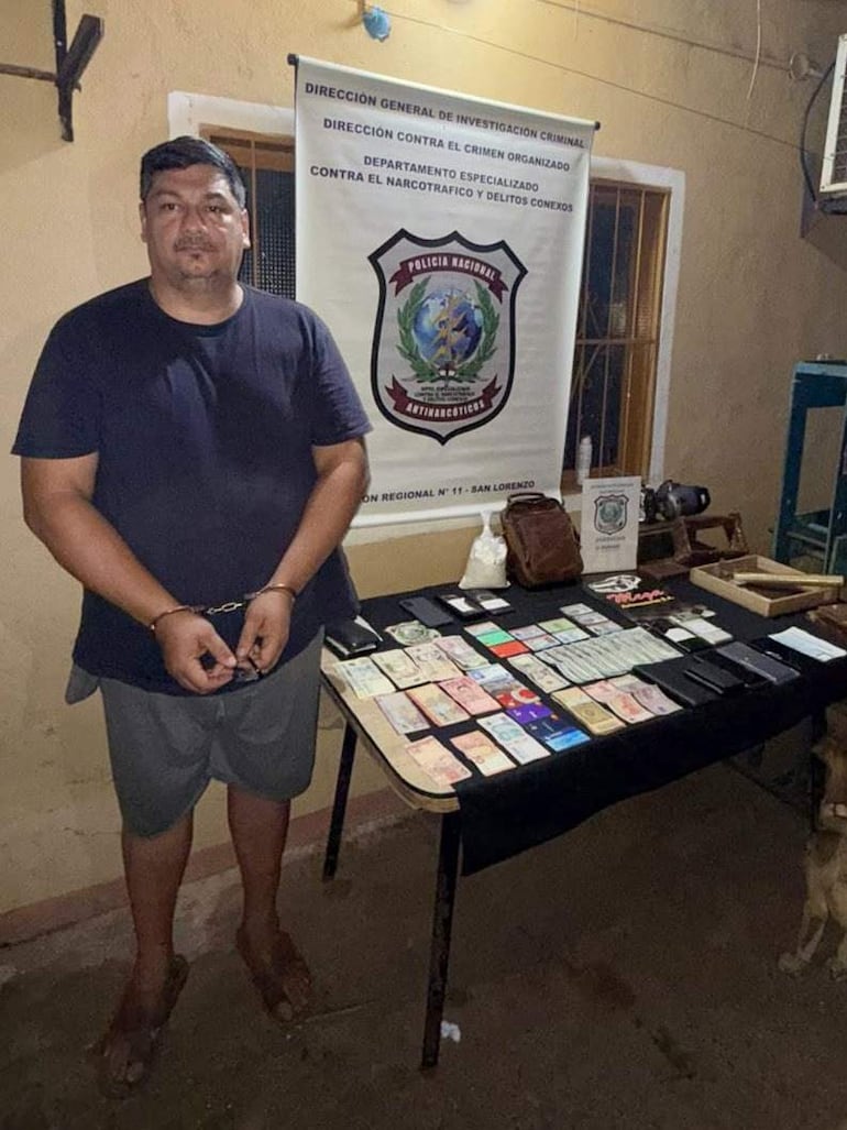 Hombre esposado con camiseta negra y shorts grises, en una sala con documentos y teléfonos, bajo la supervisión de la Policía Nacional.
