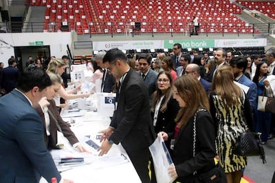 Asistentes en mesa de recepción, interactuando en ambiente organizado y formal con sillas rojas al fondo.