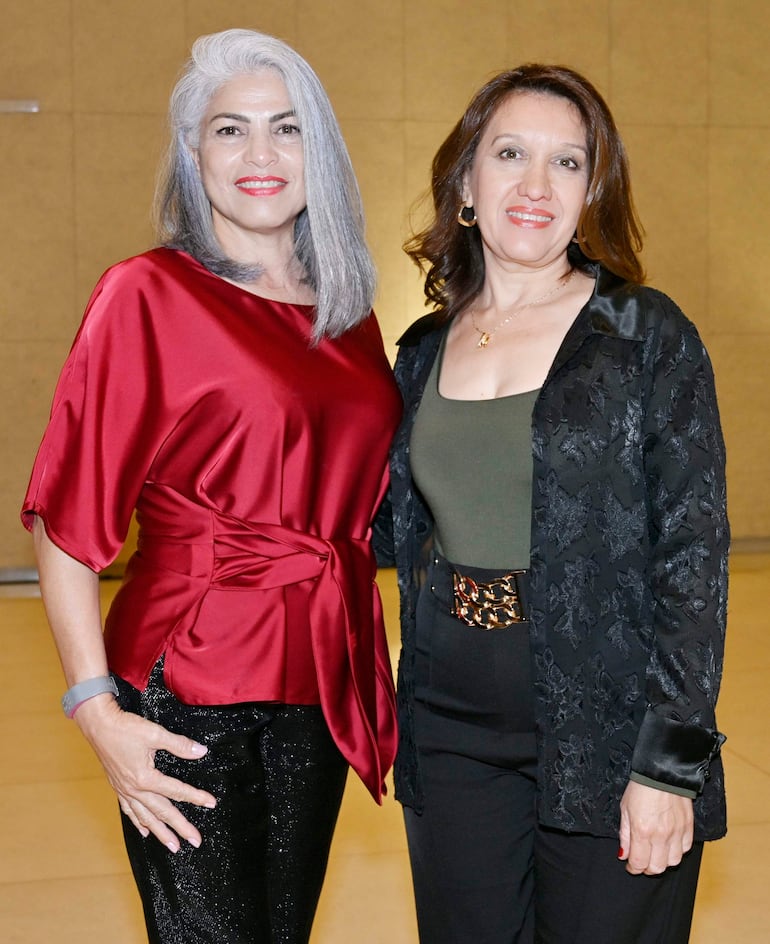 Dos mujeres sonríen, una con blusa roja satinada y otra con blusa verde y chaqueta negra, en un ambiente acogedor y elegante.