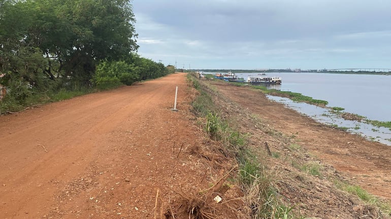Camino de tierra flanqueado por vegetación, río y barcos amarrados. Cielo nublado y ambiente rural tranquilo.