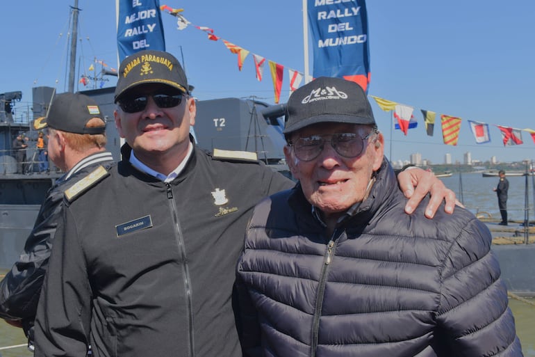 Dos hombres sonríen, uno en uniforme de la Armada Paraguaya y otro en abrigo negro, junto a una embarcación decorada y un cartel colorido.