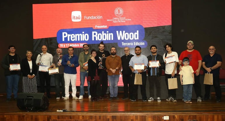 Quince premiados sonrientes en fila sosteniendo certificados, en un auditorio decorado con el logo de Itaú.