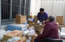 Hombre de pie en camiseta morada revisa caja, mientras otro sentado con abrigo burdeos y gafas observa con documentación en mano.
