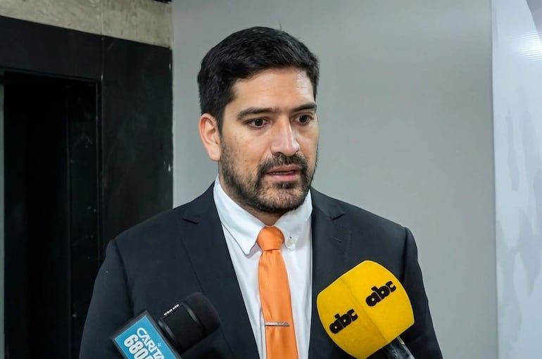 Mauricio Espínola de piel clara, con traje negro y corbata naranja, habla frente a micrófonos en un entorno interior neutro.