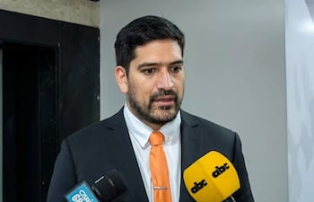Mauricio Espínola de piel clara, con traje negro y corbata naranja, habla frente a micrófonos en un entorno interior neutro.