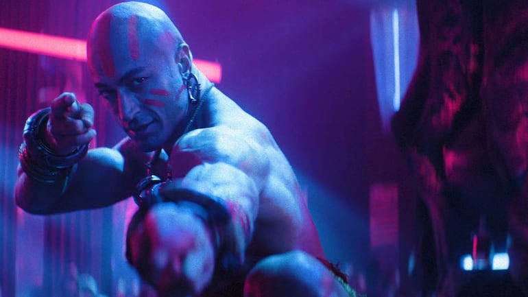 Hombre en el set de filmación, luciendo atuendo de acción, en un ambiente dinámico y de producción cinematográfica.