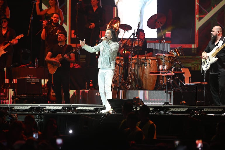 Marco Antonio Solís en chaqueta blanca actúa con micrófono, rodeado de músicos en un escenario brillante durante un concierto.