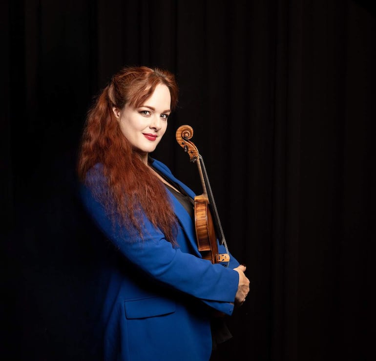Rachel Kolly de pie con mirada amigable, lleva un blazer azul y sostiene un violín en un entorno iluminado.