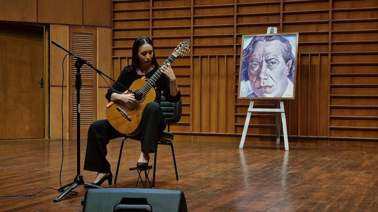 Luz Alegre sentada, tocando guitarra con expresión concentrada, en un escenario de madera, vestida de negro.