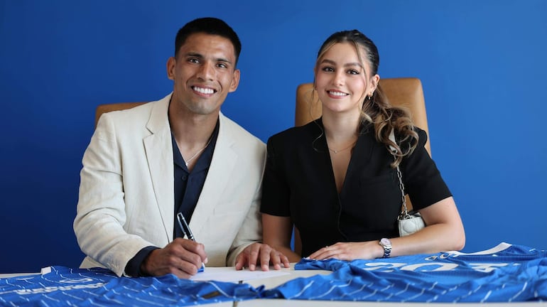 Hombre con chaqueta clara firma un documento, acompañado de una mujer vestida de negro que sonríe en fondo azul.