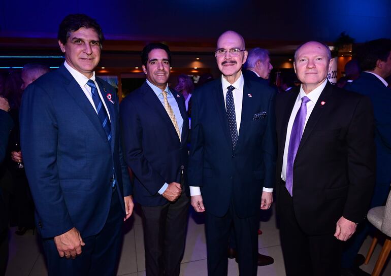 Cuatro hombres vestidos con trajes oscuros y corbatas, sonriendo en un ambiente elegante y con iluminación tenue.