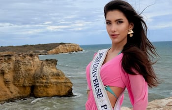Gretha Matiauda, con vestido rosa y banda de Miss Universe Paraguay 75, posa de pie en la playa con mar y cielo nublado de fondo.