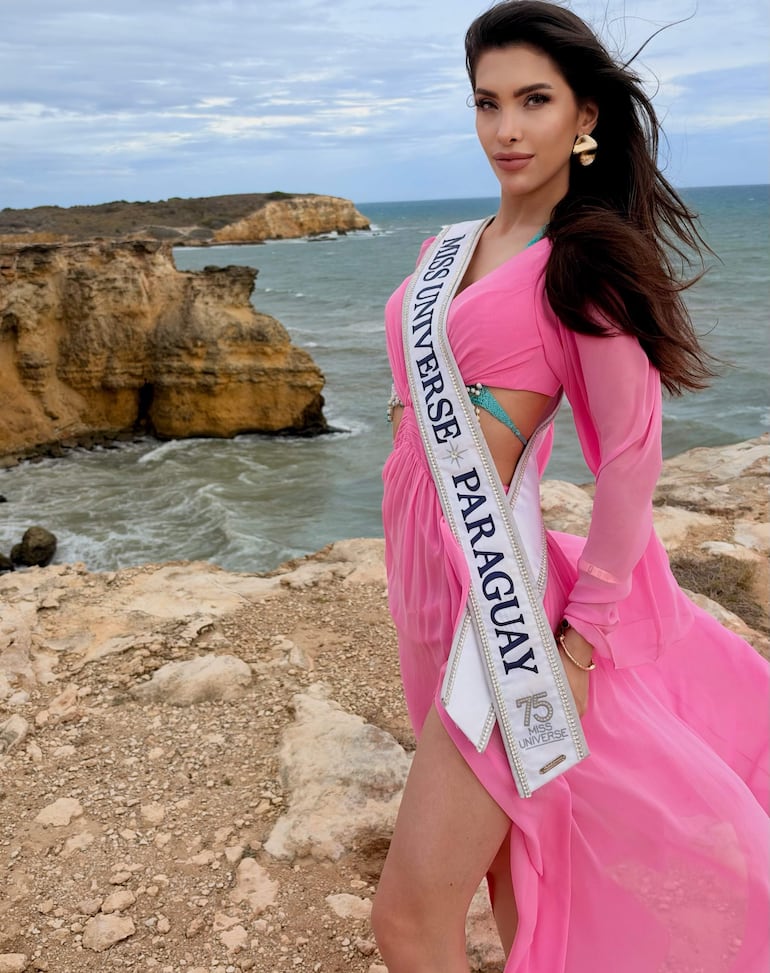 Gretha Matiauda, con vestido rosa y banda de Miss Universe Paraguay 75, posa de pie en la playa con mar y cielo nublado de fondo.