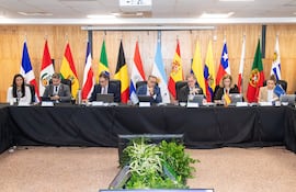 Seis individuos sentados en mesa oscura, discutiendo. Mujer a la izquierda revisa su celular, mientras el hombre central habla.