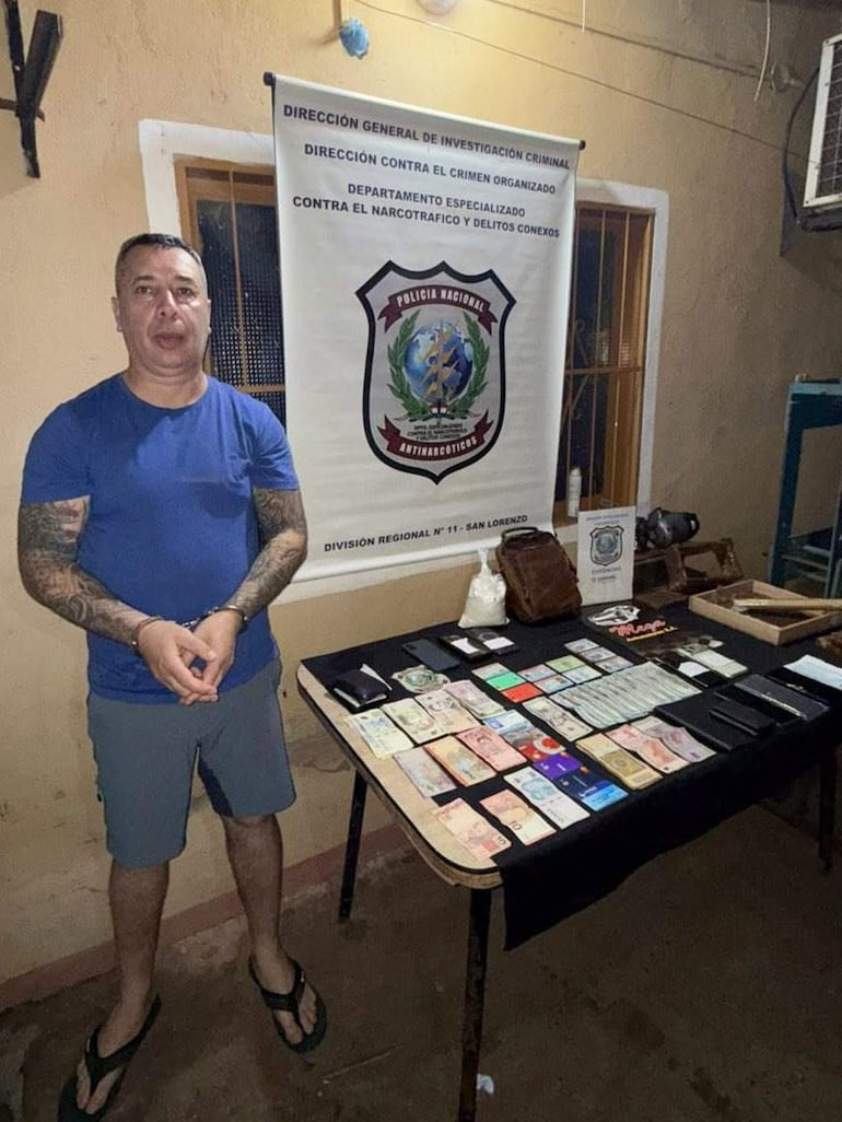 Hugo Javier Fagúndez Suárez de pie en camiseta azul y shorts, con tatuajes, frente a una mesa con objetos decomisados en una oficina policial.