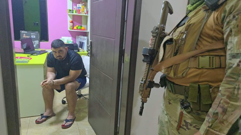 Hombre serio sentado con manos en el regazo y soldado armado en uniforme camuflado en oficina colorida.