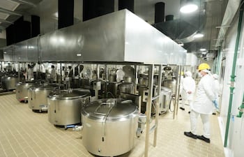 Personas en chaquetas blancas y guantes en planta de producción con recipientes metálicos y maquinaria en un ambiente industrial.