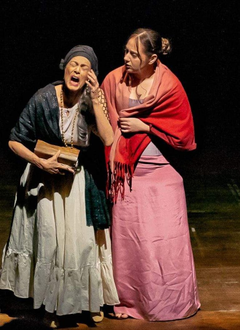 Mujer mayor con vestido blanco y chaqueta oscura, mientras la joven con vestido rosa y chal rojo la escucha atentamente en un acto teatral.