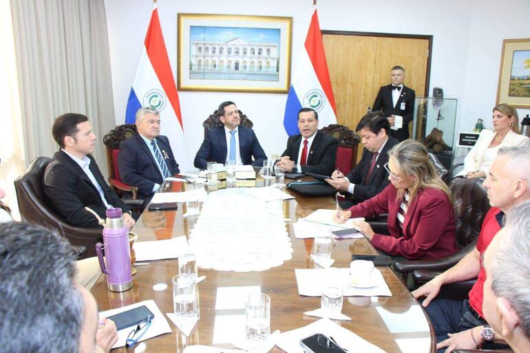 Félix Sosa y otros cinco hombres sentados alrededor de una mesa, con documentos y banderas de Paraguay al fondo, en un ambiente formal.