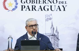 Dr. Isaías Fretes de pie frente a un micrófono, con gafas y saco oscuro, mientras presenta en el Palacio de Gobierno.