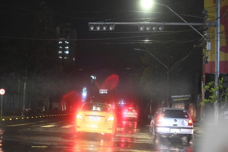 Calle urbana iluminada en la noche, con tráfico de vehículos, calles mojadas tras la lluvia.