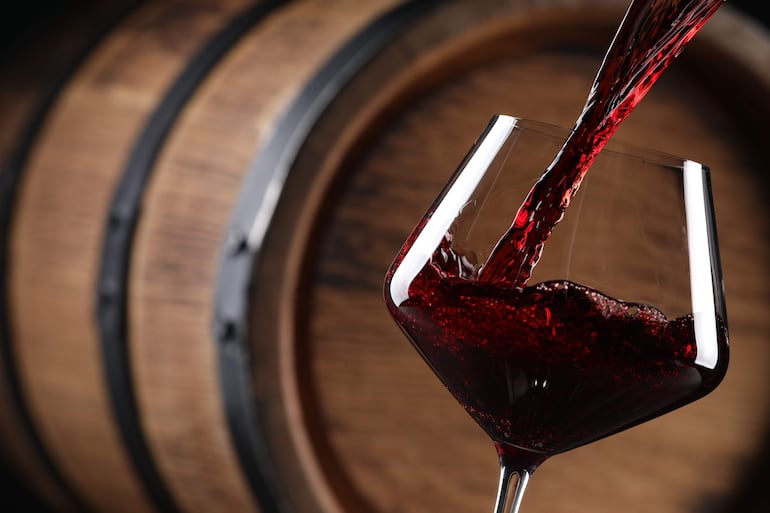 Copa de cristal con vino tinto en un entorno de bodega, barricas de madera visibles al fondo.
