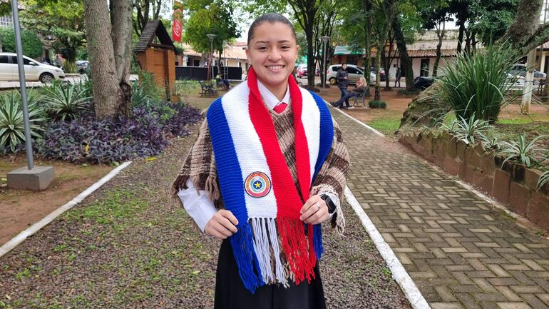 Mujer joven sonriente con bufanda tricolor y corbata roja, rodeada de árboles en festival al aire libre.