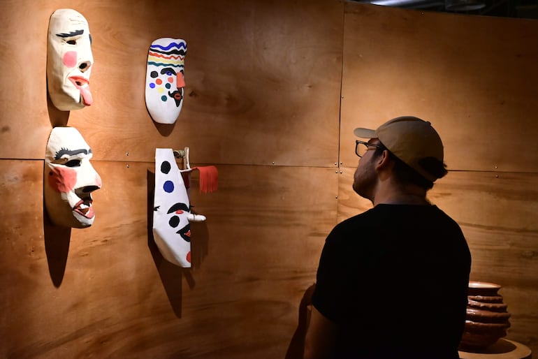 Hombre de espaldas con camiseta negra y gorra clara contempla máscaras artísticas en exposición sobre pared de madera.